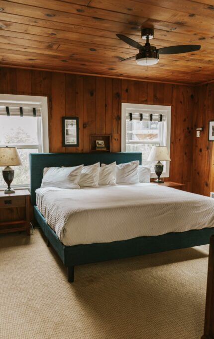 A cozy bedroom with a large bed centered against a wooden paneled wall, flanked by two nightstands with lamps. The bed has a white comforter and several white pillows. Two windows behind the bed let in natural light. A ceiling fan is installed overhead.