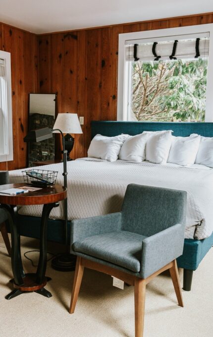 A warmly lit bedroom with wood-paneled walls featuring a large bed with five pillows and a blue headboard. Two blue cushioned chairs and a small table are in front of the bed. Two white bathrobes hang by the window. A lamp and framed photo are on the bedside table.