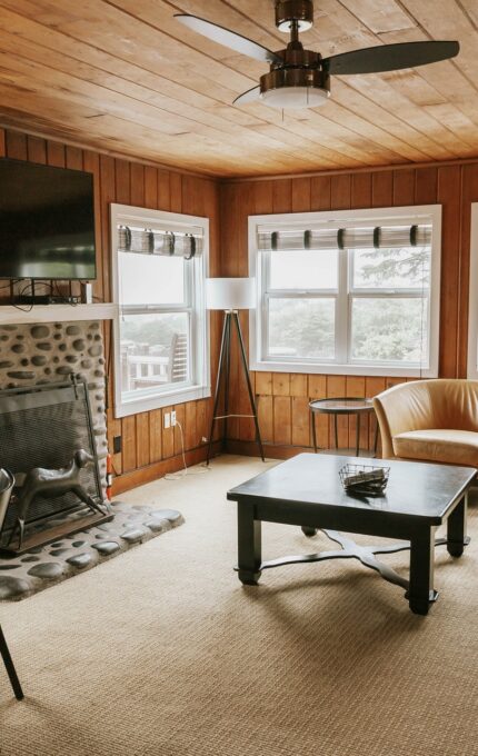 A cozy living room with wooden walls and ceiling, featuring a stone fireplace, flat-screen TV, gray sofa, round glass dining table with black chairs, armchair, and coffee table. Windows with blinds allow natural light to fill the room. Lamp and small table also present.