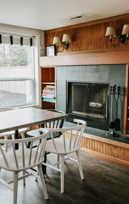 A dining area with a rectangular table surrounded by six white chairs. The area features a fireplace with a black front panel and wooden mantel, built-in shelves with books, and windows with blinds allowing natural light to enter.