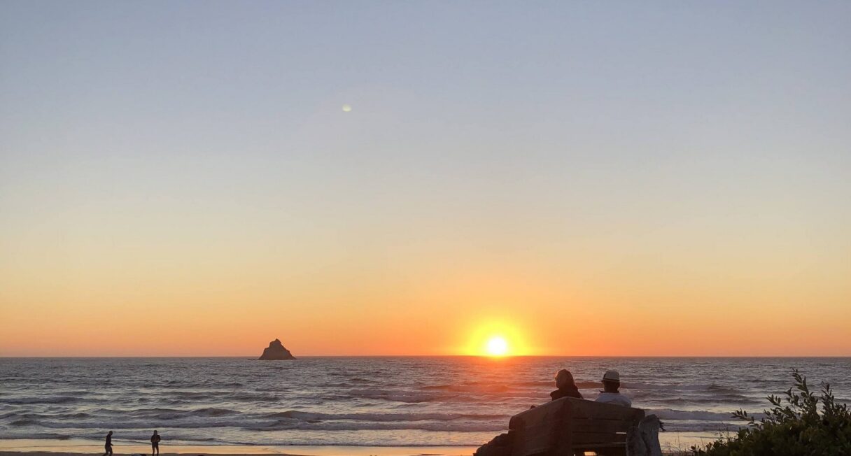 Two people sit on a wooden log at a beach, watching the sunset over the ocean. In the background, a small rocky island is visible in the distance. A few other people are seen near the water's edge, and the scene is framed by sand and scattered greenery.