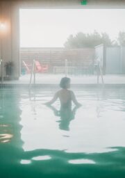 A person with short hair is standing in an indoor swimming pool, facing away from the camera. The room has soft warm lighting, and through large open windows, an outdoor seating area can be seen with red chairs and greenery in the background.