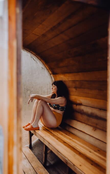 A person sits on a wooden bench inside a curved, wooden sauna. They are wearing a floral bikini top, orange bottoms, and orange slide sandals. The person has long dark hair and is gazing to the side, with a relaxed expression.