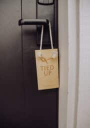 A wooden "Do Not Disturb" sign with the words "TIED UP" engraved, depicting a rope knot, hangs on the handle of a dark-colored door. The door is slightly open, showing a white wall to the right.