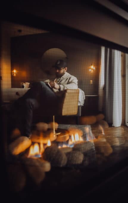 A man sits on a chair, reading a book and drinking from a mug in a dimly lit room. A lit fireplace with visible flames is in the foreground. He wears a cap and appears relaxed in the cozy setting.