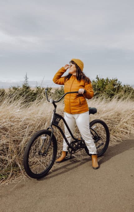 A person stands next to a black bicycle on a path. They are wearing a mustard yellow jacket, matching beanie, white pants, and brown boots. The person holds their right hand up to shield their eyes while surrounded by tall grass with an overcast sky in the background.