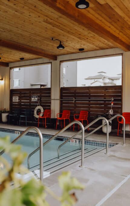 An indoor pool with metal handrails on the sides and tiled edges. A wooden ceiling and modern light fixtures are visible above. There are red chairs arranged along a wooden fence with life rings hanging on it in the background. Some buildings are visible through open windows.