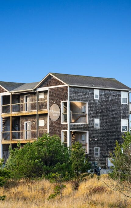 A three-story wooden house with balconies on each floor is situated in a grassy area surrounded by trees. The exterior is clad in dark shingles. The sky overhead is clear and blue.