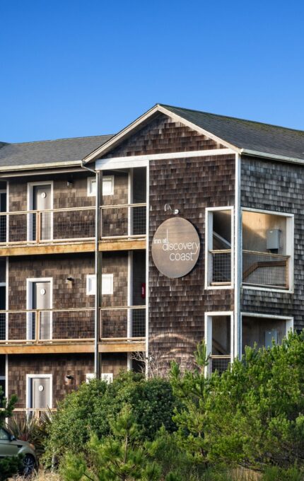 A three-story, brown-shingled apartment building with balconies is partially obscured by trees. A round sign on the front reads "Discovery Coast." The building stands against a clear blue sky.