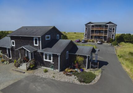 A two-story house with a wooden exterior stands next to a three-story apartment building. The area features paved driveways, green grassy surroundings, and a few parked cars. The sky is clear with a hint of fog in the background.