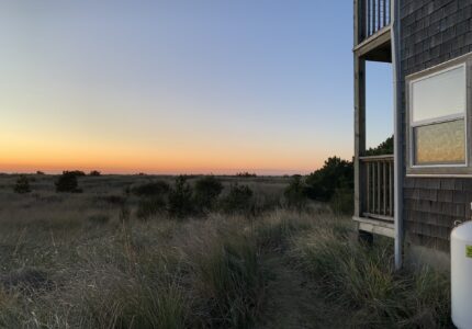 A two-story building stands on the right side of an outdoor scene overlooking a vast grassy field at sunset. The sky transitions from a golden hue near the horizon to a deep blue at the top.