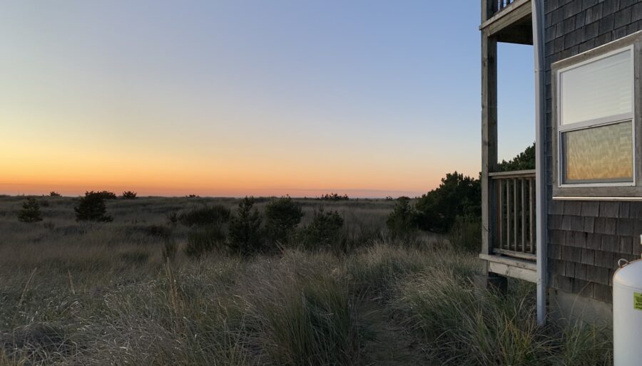 A two-story building stands on the right side of an outdoor scene overlooking a vast grassy field at sunset. The sky transitions from a golden hue near the horizon to a deep blue at the top.
