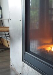 A close-up of a lit fireplace with logs burning behind a glass screen. In the background, there is a small table with two wicker chairs, a bottle of wine, and two wine glasses set on the table. The scene captures a cozy indoor setting.