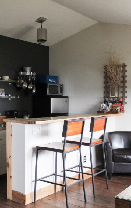 A small kitchen area with white cabinetry, a microwave, and a coffee maker. Two wooden barstools are placed at the counter. In the adjacent living area, there are two leather chairs, a wall-mounted black and white picture, and a wooden chest used as a table.