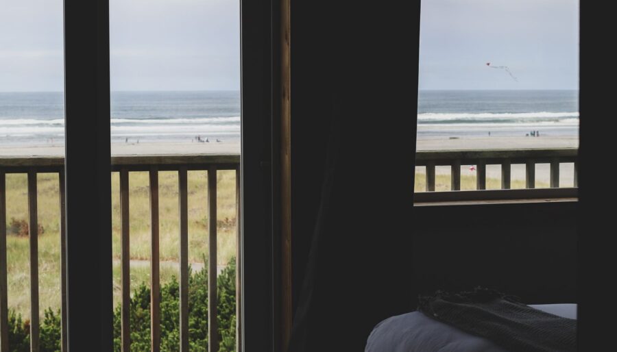 View of a beach from a room with wooden balcony doors. The sandy shore and ocean waves are visible through the open doors, while part of a bed with a blanket is seen in the foreground. A kite flies in the sky over the beach.