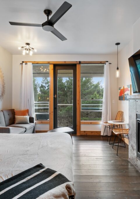 A cozy modern bedroom features a large bed, a sofa, a fireplace, and a wall-mounted TV. There's a small dining area with two chairs and a table. The room has a balcony with a view of trees. The decor is minimalistic with neutral tones and wooden accents.