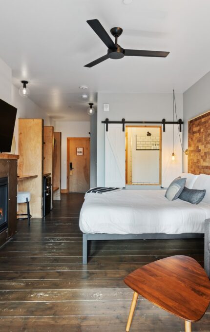 A modern hotel room with a bed, gray sofa, wooden coffee table, and a wall-mounted TV above a fireplace. The room features a wooden floor, ceiling fan, wicker chair, and a sliding barn door. Lighting includes pendant lights and natural light from a window.