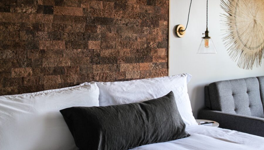 A neatly made bed with white sheets and pillows, and a black accent pillow, is in front of a textured brown wall. A pendant light with an exposed bulb hangs on the right, next to a gray upholstered couch and a decorative wall hanging.