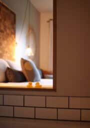 A softly lit bedroom with a view through an open window frame. The bed has pillows and a blanket. Two small yellow rubber ducks sit on a tiled ledge in the foreground. The background includes a wall with hanging lights and art.