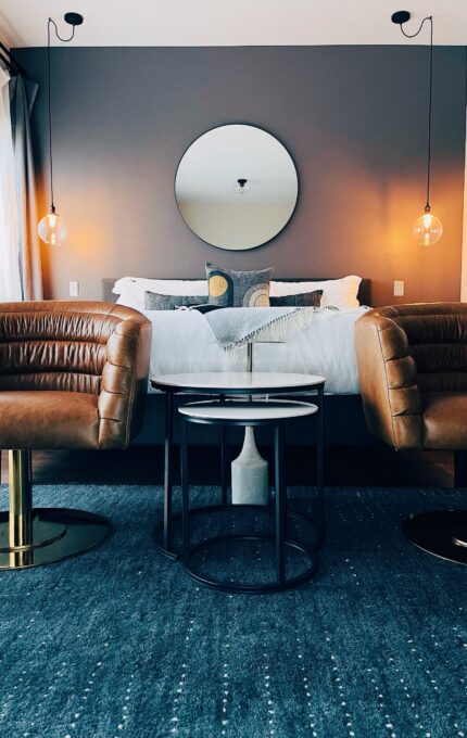 A modern bedroom features a neatly made bed against a dark accent wall with a round mirror above it. Two hanging pendant lights flank the bed. In the foreground, two brown leather chairs face a pair of nested side tables on a blue rug.