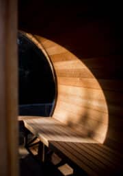 A wooden outdoor sauna with curved walls and a large round window partially illuminated by sunlight. The interior features wooden benches on either side. The light creates a sharp contrast between the bright and shaded sections.