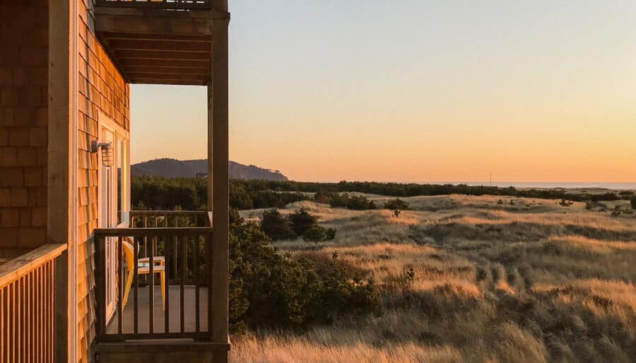 A wooden building with several balconies overlooks a landscape of dry grass and distant hills under a clear sky during sunset. Each balcony has seating, and the sunlight casts a warm glow over the scene.