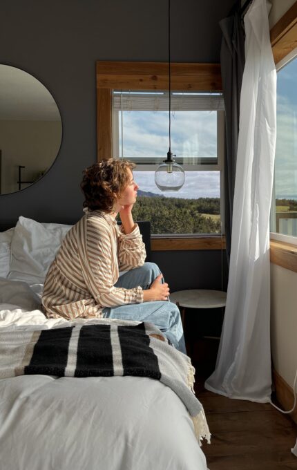 A person with short hair is sitting on a bed by a window, looking outside. They are wearing a striped shirt and jeans. The room has a dark gray wall, a round mirror, and a hanging light bulb. The bed is unmade with a striped blanket at the foot.