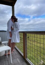 Two people are standing on a wooden deck with a wire railing, overlooking a grassy field under a cloudy sky. They are both wearing white bathrobes and are standing close together. A small table with a few items on it is visible to the left.