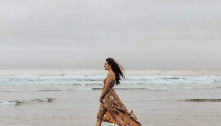 A woman in a patterned dress walks barefoot along a beach with waves in the background. The sky is overcast.