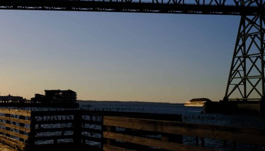 A view of a waterfront with a large steel bridge overhead during sunset. The pier below is made of wood, extending towards the water, with buildings and the horizon visible in the background.