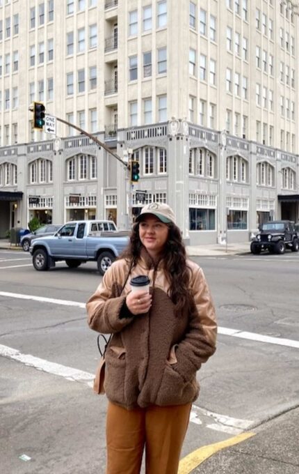 A person stands at a street corner in front of a multi-story building, holding a cup of coffee. The person is wearing a brown jacket, tan pants, and a gray cap. Several vehicles are stopped at the intersection. The building has a vintage architectural style.