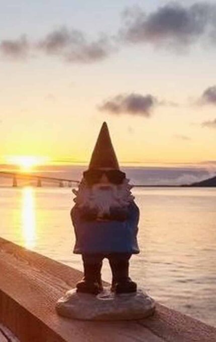 A gnome figurine with sunglasses stands on a railing by a waterfront at sunrise. The scene includes a bridge in the background and clouds in the sky reflecting the early morning light.