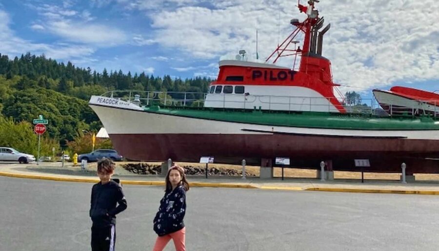 Two children stand with arms raised in front of an old, red and white pilot boat on land. The boat is named "PEACOCK" and is situated near a wooded area under a partly cloudy sky.