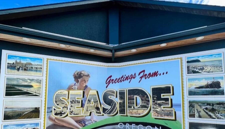 A mural on a building depicts a vintage-style postcard with the text "Greetings From... SEASIDE OREGON." The mural features images of the coastline, beach scenes, and a smiling woman in retro attire. The sky above is blue with scattered clouds.