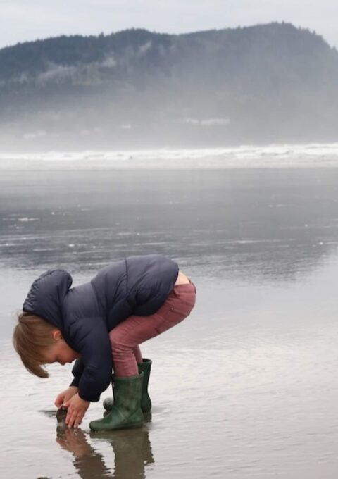 A child in a dark jacket and green boots crouches down to inspect something on a wet, reflective beach with a backdrop of foggy mountains and ocean waves.