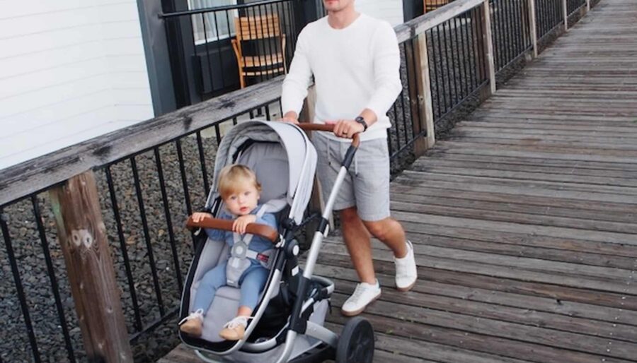 A man pushes a baby in a stroller along a wooden boardwalk. They are near a building with balconies and glass doors. The baby is seated in the stroller, wearing a blue outfit and looking forward, while the man is wearing a white shirt and gray shorts.