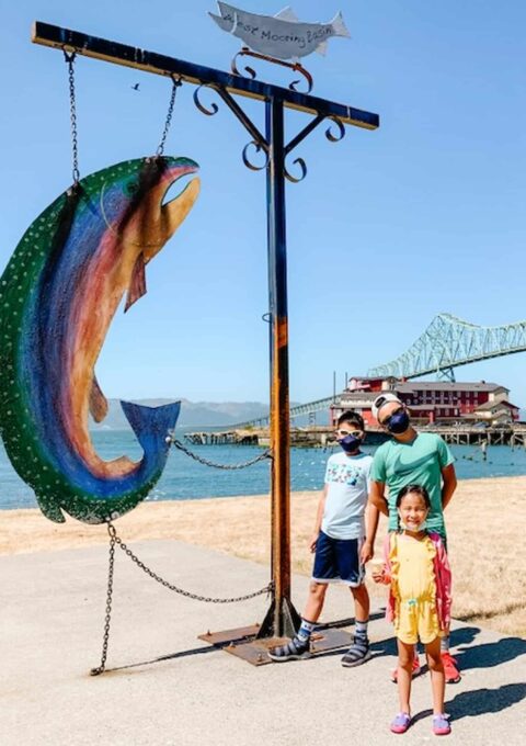 A family stands next to a large colorful fish sculpture outdoors. The father and two children are all wearing casual clothing and face masks. A bridge and body of water are visible in the background under a clear blue sky.