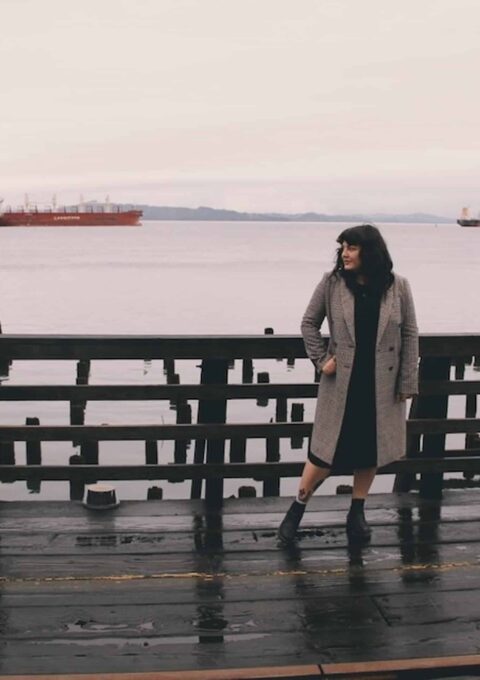 A person wearing a long coat and boots stands on a wet wooden pier gazing to the side. In the background, two red cargo ships are anchored on a calm body of water under an overcast sky. The scene has a muted color palette, indicative of a cloudy day.