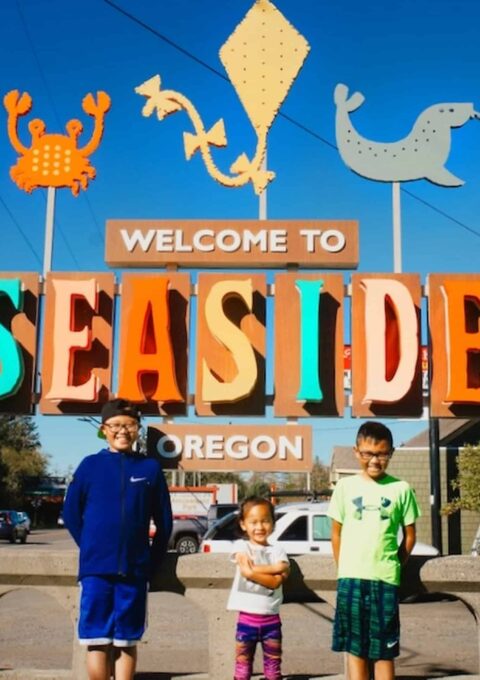 Three children stand smiling in front of a colorful "Welcome to Seaside, Oregon" sign. The sign features colorful artwork of a crab, starfish, and fish. The scene is outdoors with a clear blue sky.