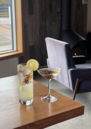 A wooden table with two drinks: a tall glass with a lemon slice and herbs next to a martini glass with similar garnish. A window and a modern fireplace with a chair are in the background.