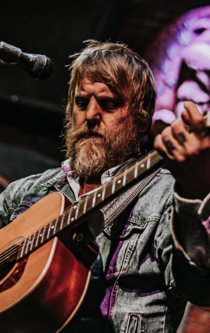 A man with a beard plays an acoustic guitar on stage. He is wearing a denim jacket and stands in front of a microphone. A circular decorative sign is visible in the background on a brick wall.