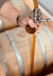 A person is using a nozzle attached to a hose to fill a wooden barrel with liquid. The hand of the person is holding the nozzle close to the top of the barrel. The barrels are made of light wood with metal bands.