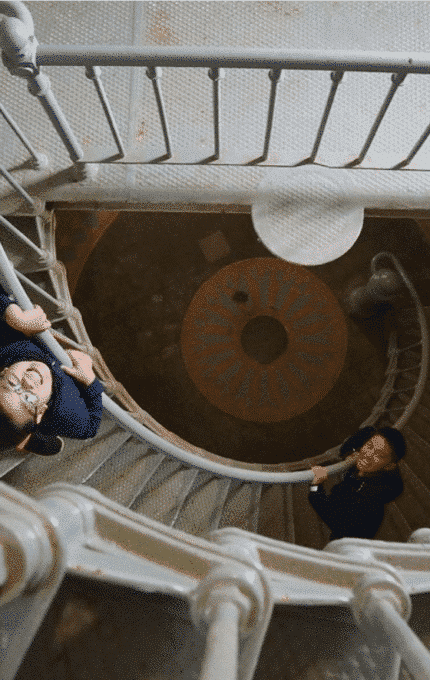 Two children are ascending a spiral staircase, looking up towards the camera. One child grips the railing, smiling, while the other stands a few steps below. The staircase is metal with several flights visible beneath them.