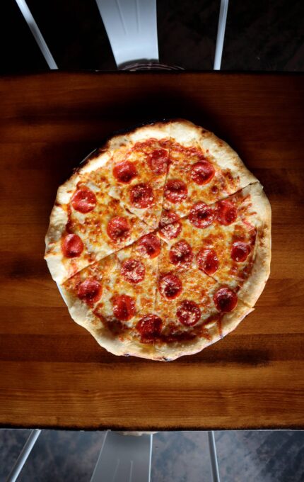 A pepperoni pizza with melted cheese is centered on a wooden table. The pizza is divided into eight slices, with one slice slightly pulled away from the rest. The crust is golden brown and the pepperoni slices are evenly distributed.