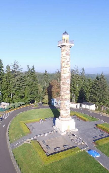 Aerial view of Astoria Column, a tall, cylindrical tower standing in a clearing surrounded by trees and a parking area. The column is decorated with a spiral mural. The scene includes parked vehicles and a backdrop of forested hills under a clear sky.