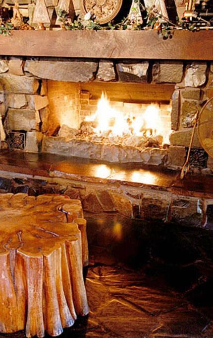 A cozy living room with a large stone fireplace actively burning. A wooden stump coffee table is placed in front of a green armchair. Decorative items, including a metal shield and various ornaments, are arranged on the mantel and hearth.