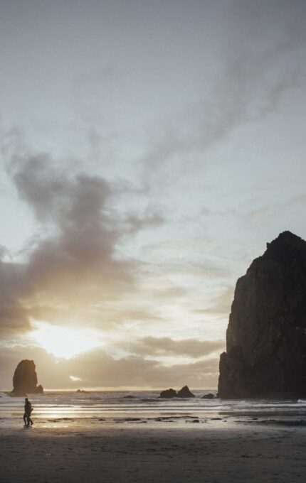 A coastal scene at sunset features large rock formations rising from the ocean. Several people are visible walking along the shoreline, and the sky is partially covered in clouds, allowing sunlight to stream through, reflecting on the water.