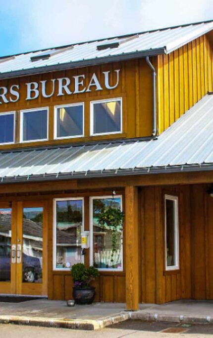 A wooden building with a metal roof labeled "Visitors Bureau" featuring large glass windows and double doors. Two teal Adirondack chairs are positioned under the right front window. Potted plants decorate the entrance area.