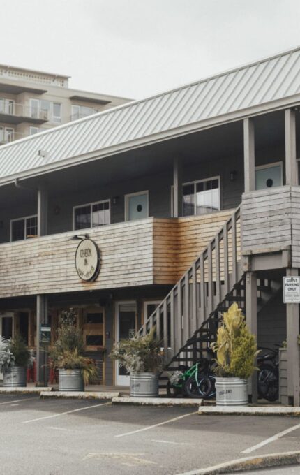 A two-story motel with wooden siding and a metal roof. The motel sign reads "Ashore Hotel." The building has exterior staircases and doors leading to each room. The surroundings include potted plants and a parking area. A building labeled "Seaside" is visible in the background.