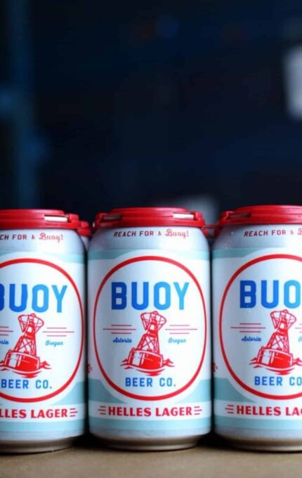 Three cans of Buoy Beer Co. Helles Lager are positioned side by side on a surface. The cans have a white background with a red and blue design, featuring a buoy illustration and text stating "Astoria, Oregon.